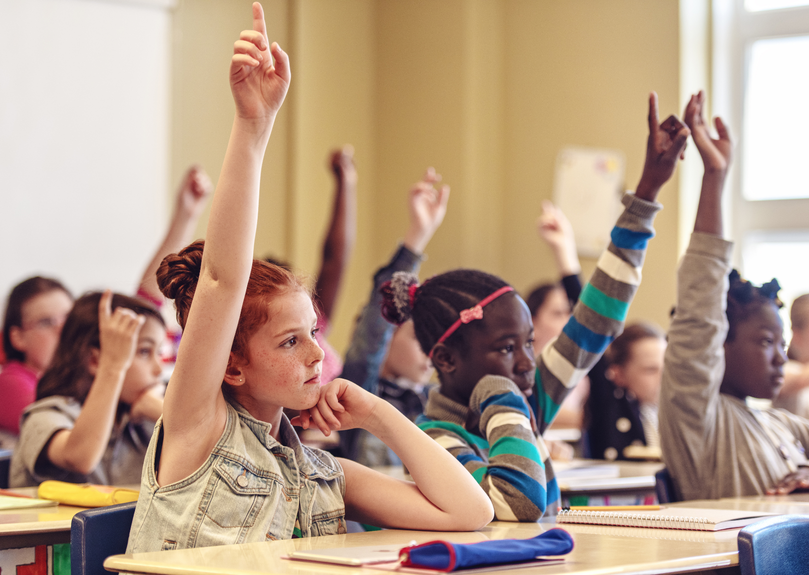 Students in a classroom raise their hands to answer a teacher's question, demonstrating growth in their knowledge.